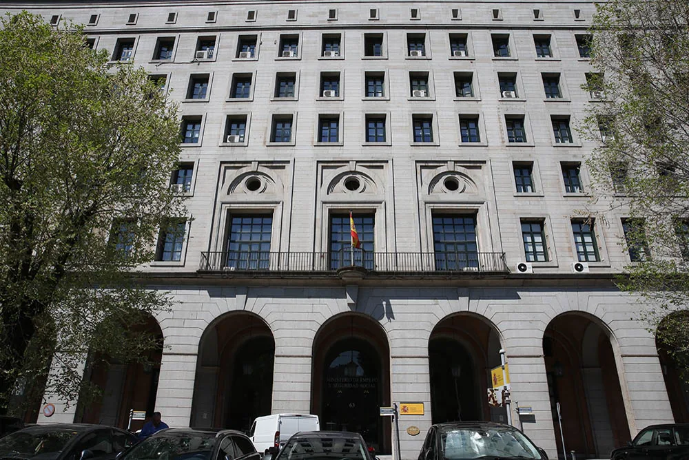 Fachada de piedra del edificio del Ministerio de Trabajo y Economía Social, en Nuevos Ministerios (Madrid), con la bandera de España en el balcón central, soportales en la planta baja y coches aparcados delante.