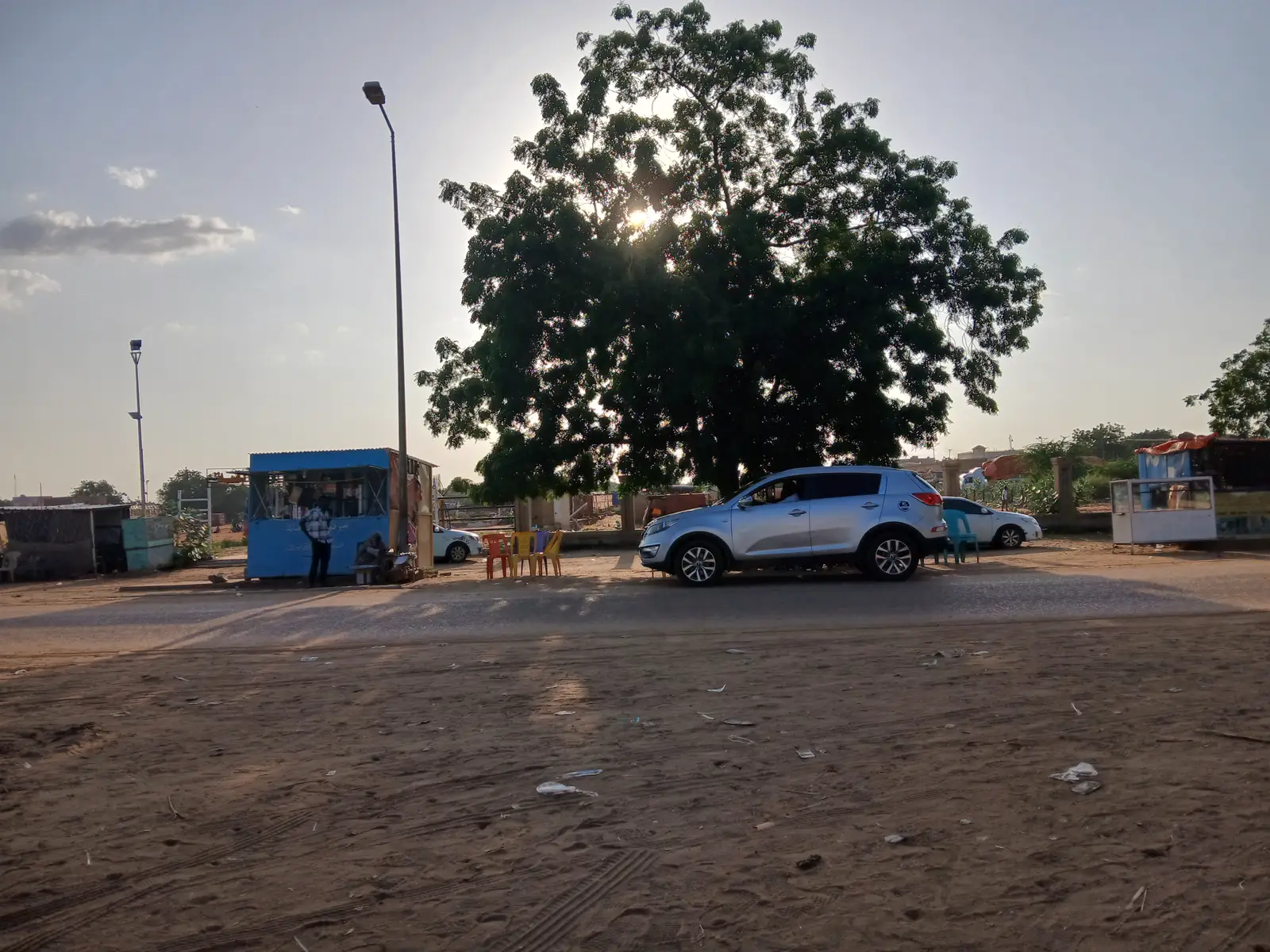 Calle de arena de El Obeid al atardecer, con un gran árbol, una caseta azul, sillas de plástico y varios coches aparcados.