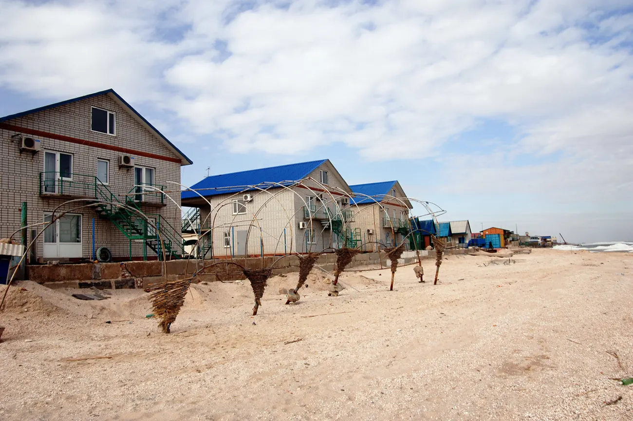 Hilera de casas de dos plantas con tejados azules levantadas al borde de una playa de arena clara, con el mar a la derecha y el cielo nublado.