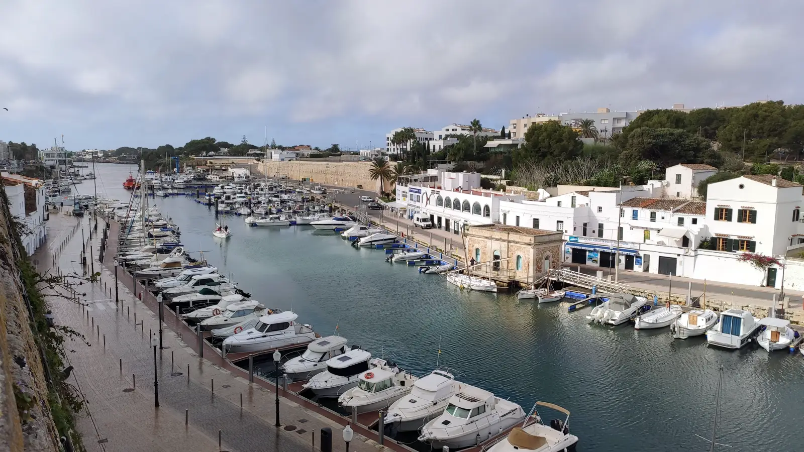 Vista elevada del puerto de Ciutadella de Menorca: un canal de agua estrecho y alargado entre dos muelles, con decenas de embarcaciones de recreo amarradas a ambos lados y casas blancas al fondo.