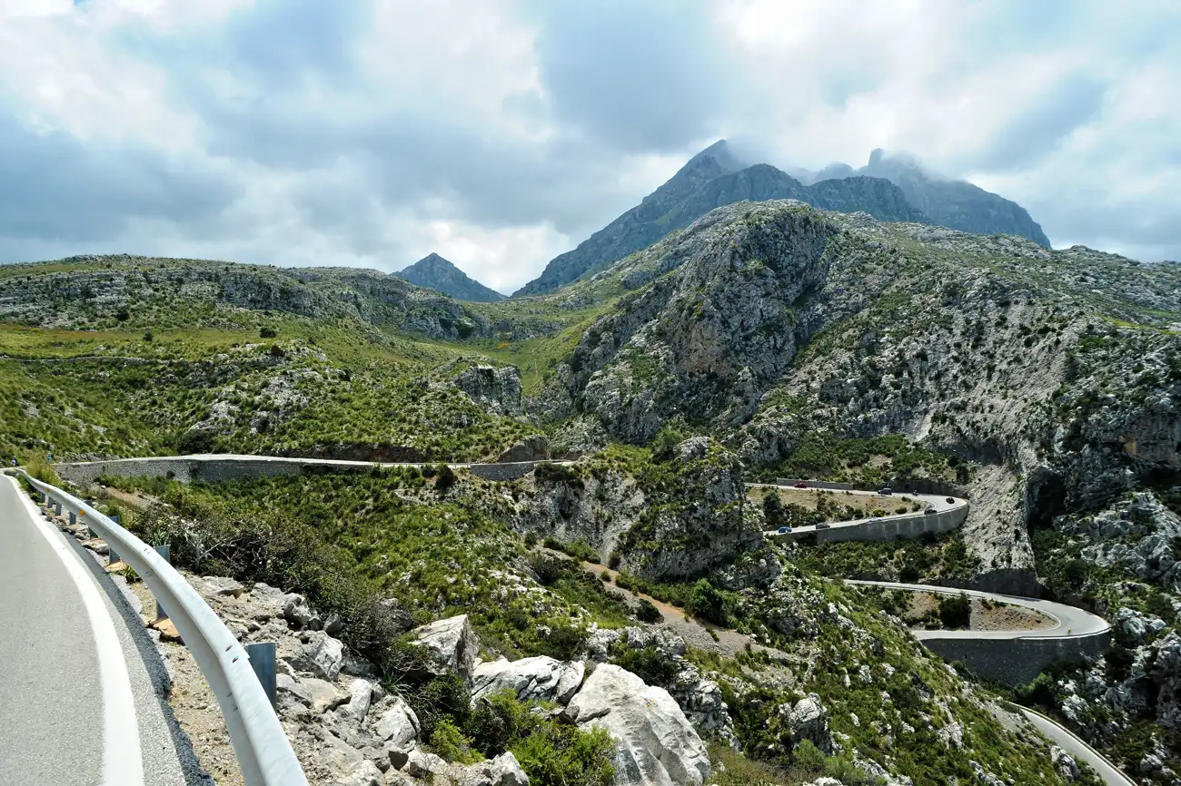 Panorámica de la Serra de Tramuntana desde la carretera de Sa Calobra, en Mallorca: laderas de roca caliza y matorral con la carretera trazando curvas cerradas en primer plano y las cumbres del fondo tapadas por nubes bajas y oscuras.