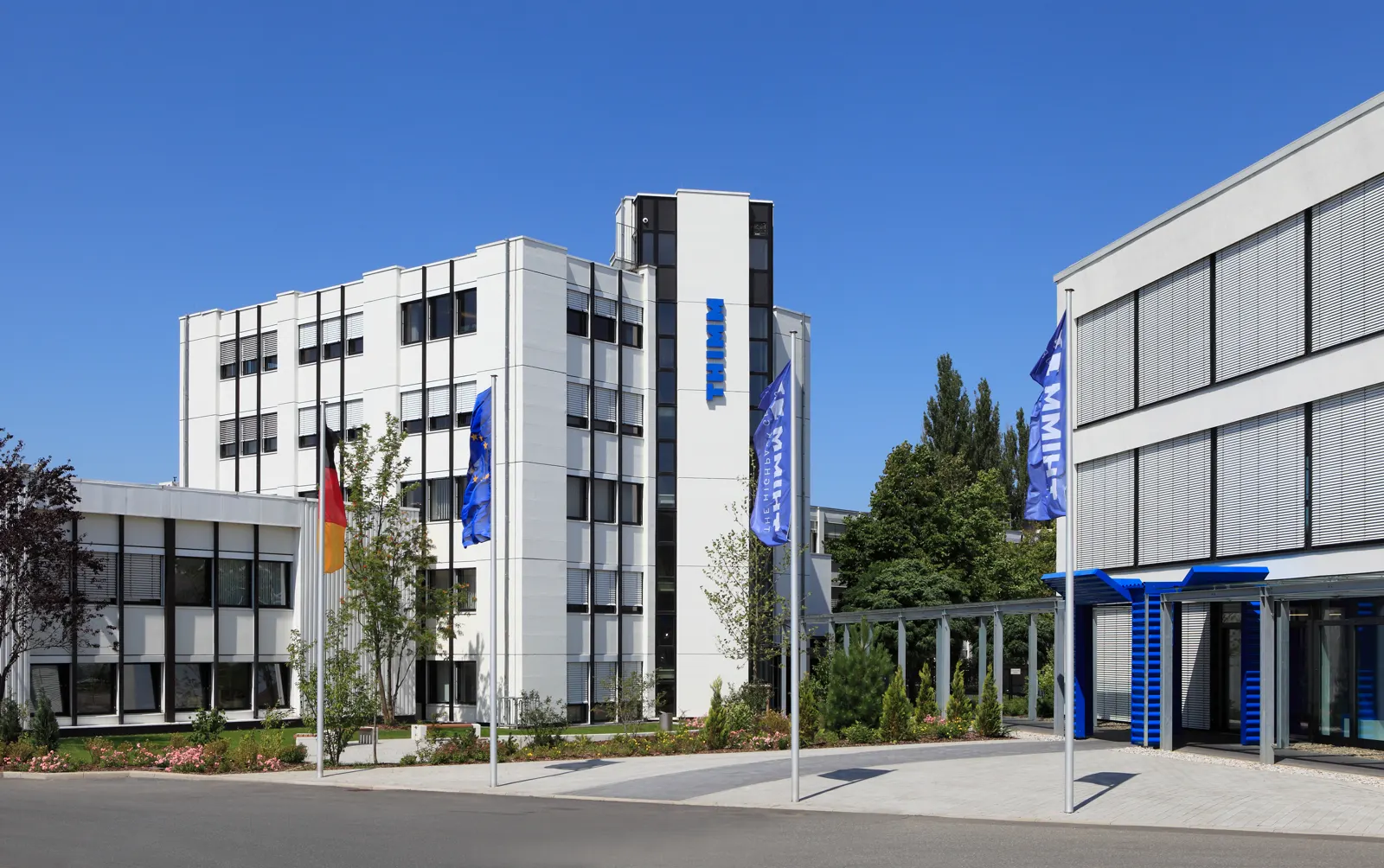 Edificios blancos de oficinas de la sede central de THIMM, con el rótulo azul de la empresa en la fachada y banderas de Alemania, la Unión Europea y THIMM en el acceso