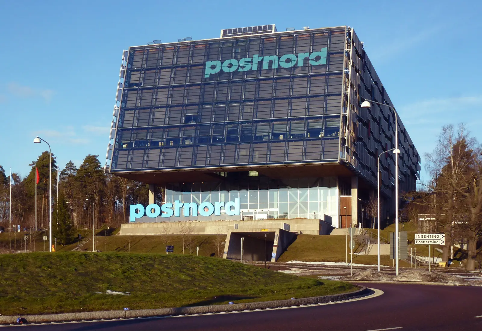 Edificio acristalado de varias plantas, en voladizo sobre una base de cristal, con el logotipo «postnord» en letras turquesa en la fachada y delante de la entrada, bajo un cielo azul y con nieve en el suelo.