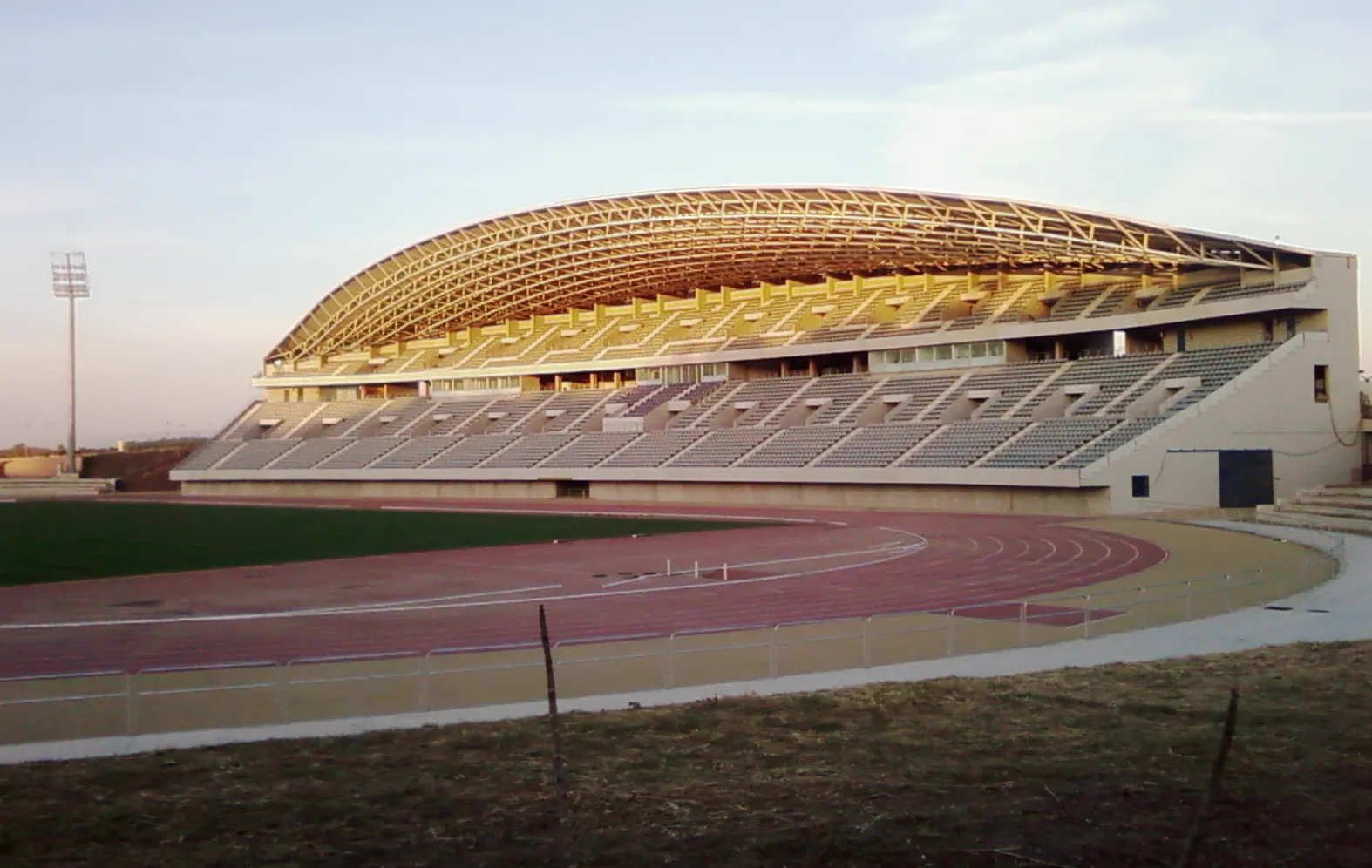 Grada principal cubierta y pista de atletismo del Estadio Ciudad de Málaga, vacío, con el tartán rojo en primer plano.