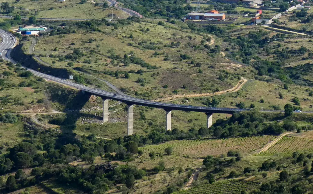 Vista aérea de un viaducto de carretera sostenido por cuatro pilas de hormigón que cruza un valle de laderas verdes, con viñedos, encinas, casas y naves industriales al fondo.