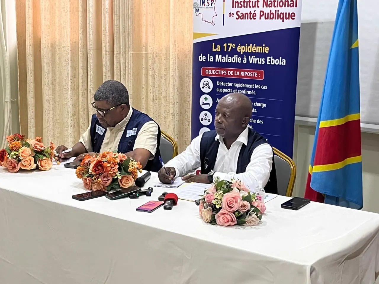 Dos responsables sanitarios sentados a una mesa con micrófonos y cuadernos, ante un cartel del Instituto Nacional de Salud Pública sobre la 17.ª epidemia de ébola y la bandera de la República Democrática del Congo