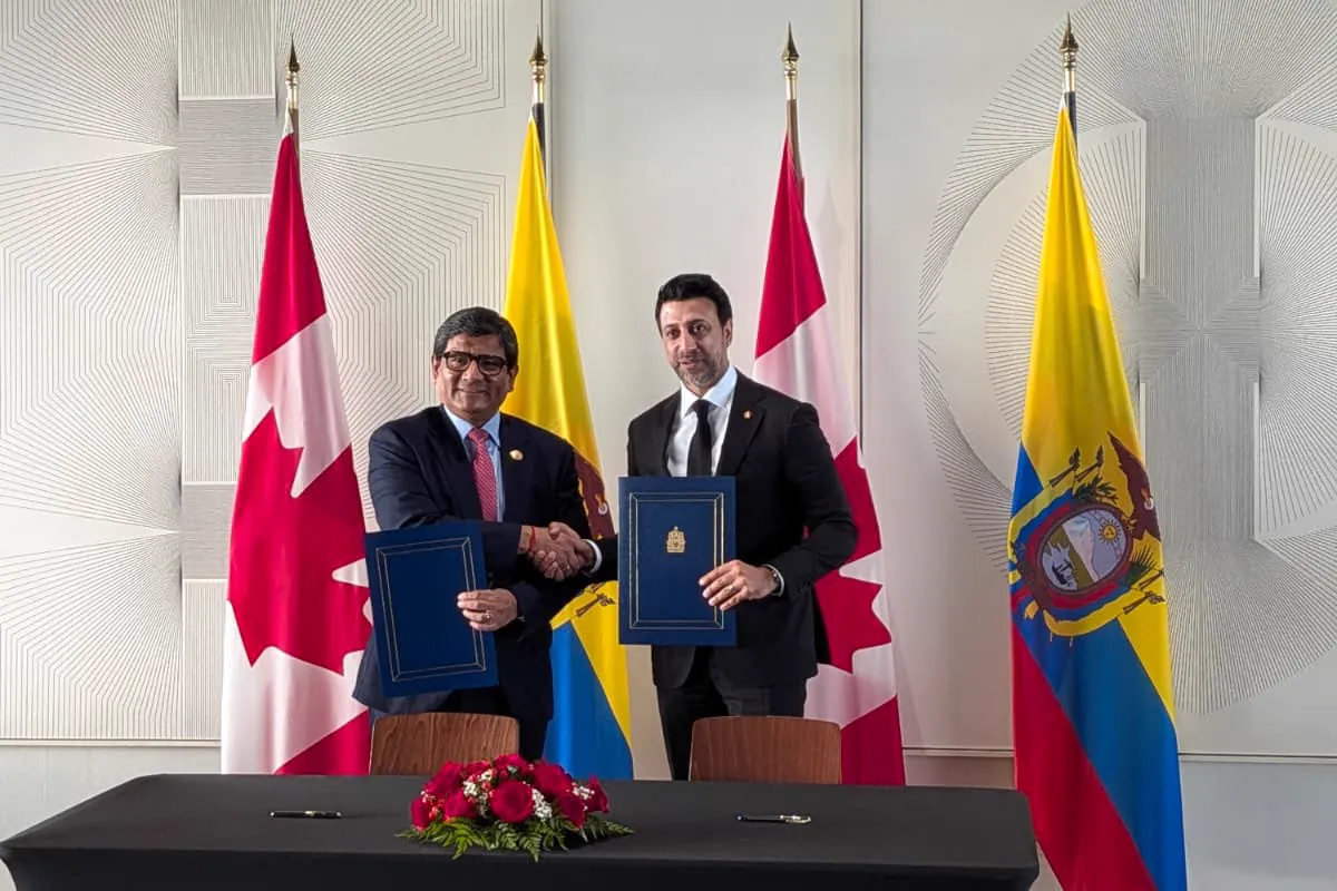 Dos hombres trajeados se estrechan la mano sosteniendo sendas carpetas azules con un documento firmado, ante banderas de Canadá y de Ecuador y una mesa negra con dos bolígrafos y un centro de rosas rojas.