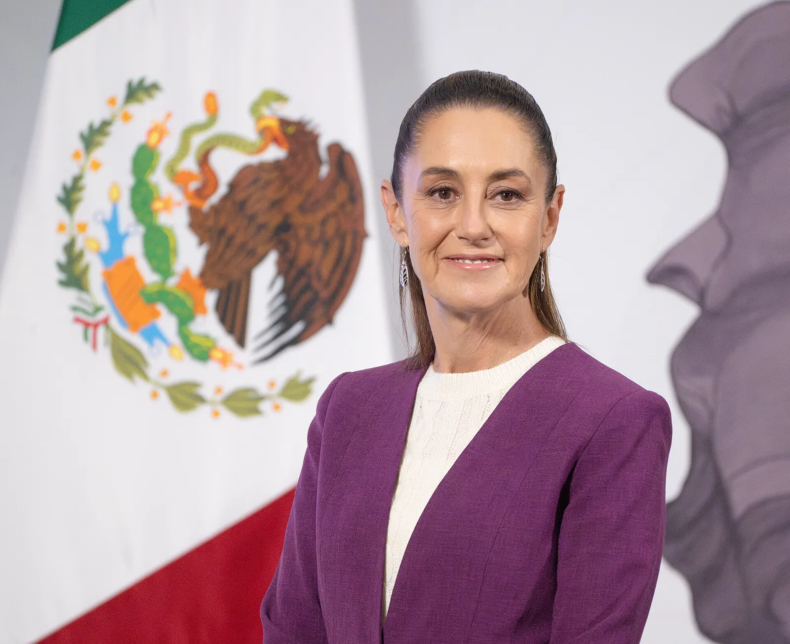 La presidenta de México, Claudia Sheinbaum, sonríe ante la bandera mexicana durante una conferencia de prensa.