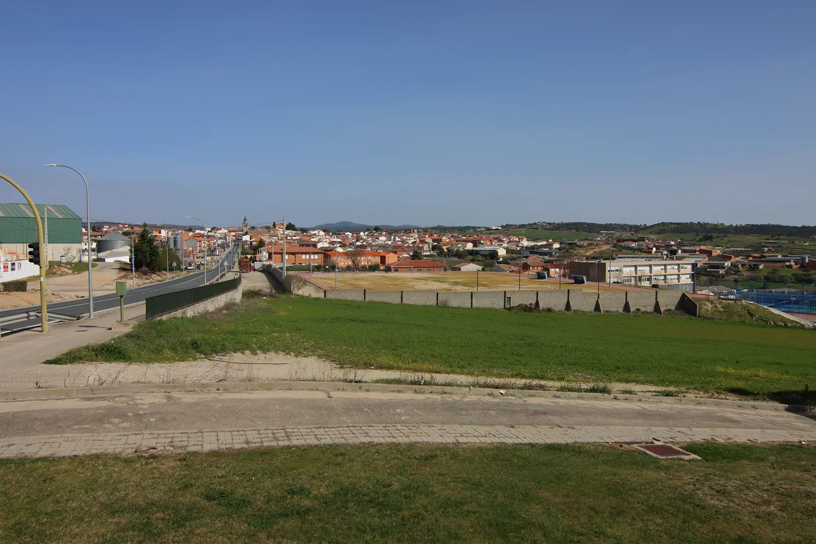 Vista del casco urbano de Almorox (Toledo) desde la carretera N-403, que baja por la izquierda de la imagen entre farolas y una nave industrial; entre las casas destaca la torre de la iglesia y al fondo se ven lomas de monte bajo bajo un cielo despejado.