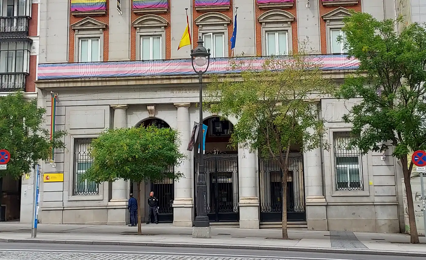 Fachada de piedra de un edificio institucional en una calle de Madrid, con una entrada porticada entre columnas, banderas de España y de la Unión Europea, banderolas de colores en el primer piso y dos personas junto a la puerta.