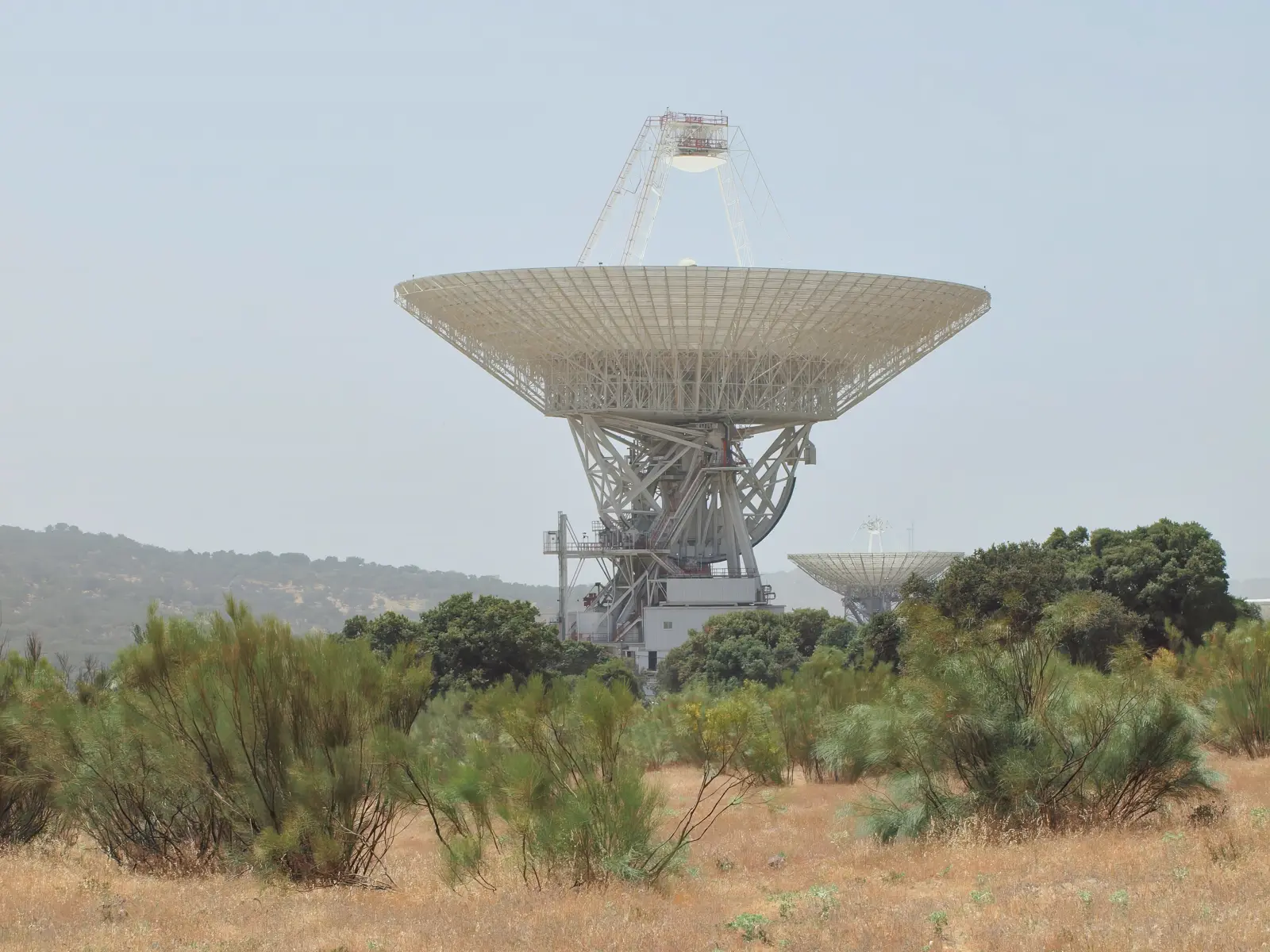 Gran antena parabólica blanca sobre una estructura metálica, con otra antena menor al fondo, rodeada de pasto seco y matorral bajo en una loma de monte mediterráneo.