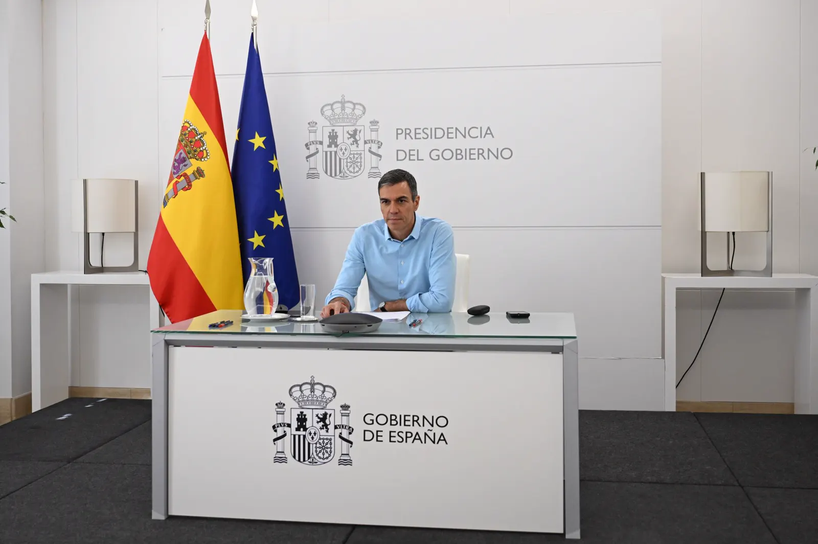 Pedro Sánchez, con camisa azul claro, sentado solo ante una mesa de cristal con un micrófono de videoconferencia, una jarra de agua y unos papeles, en una sala blanca con las banderas de España y de la Unión Europea y el rótulo «Presidencia del Gobierno» en la pared.