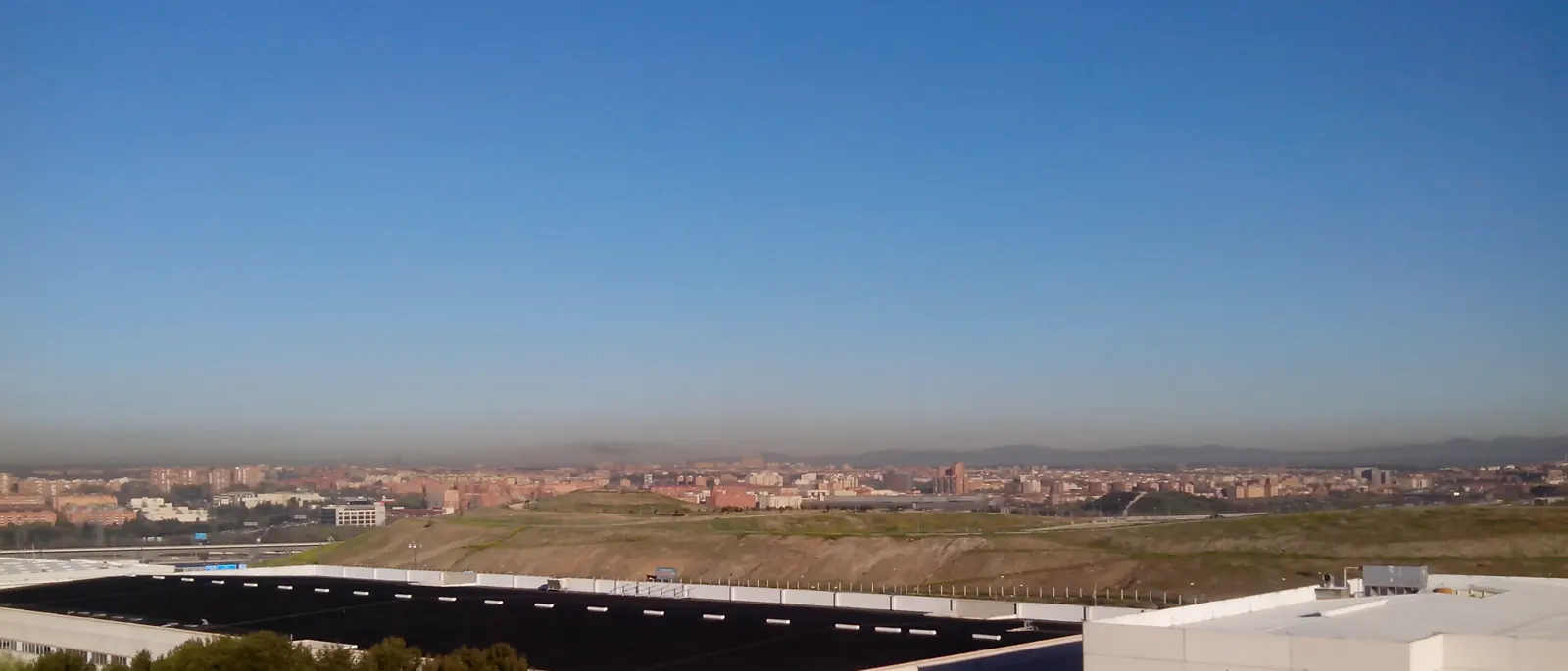 Vista panorámica de Madrid desde una loma del extrarradio: bajo un cielo azul, una franja de aire pardo y turbio se extiende de lado a lado sobre los edificios de la ciudad, con la sierra recortada al fondo.