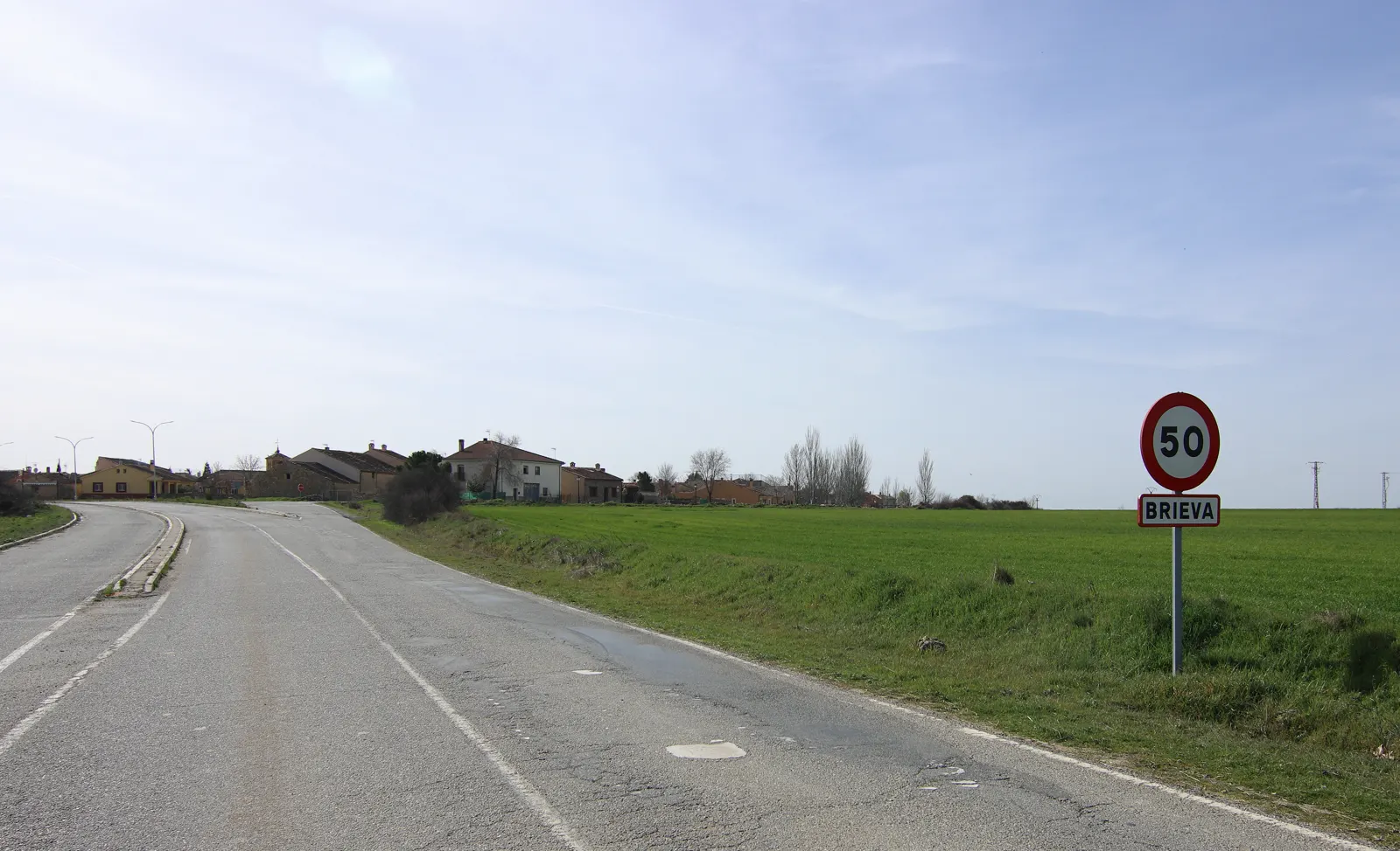 Carretera de acceso a Brieva (Segovia) con la señal de entrada al pueblo a la derecha, campos verdes a ambos lados y las casas de la localidad al fondo.