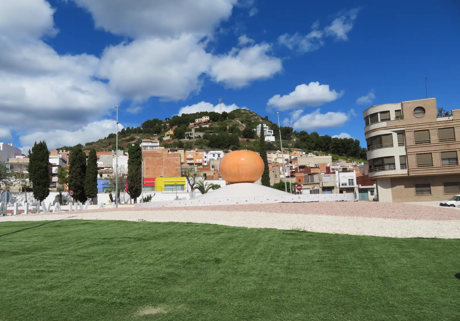 Rotonda de entrada a La Vilavella con una escultura de una naranja gigante y las letras del nombre del municipio; detrás, las casas del pueblo y un cerro cubierto de pinar.