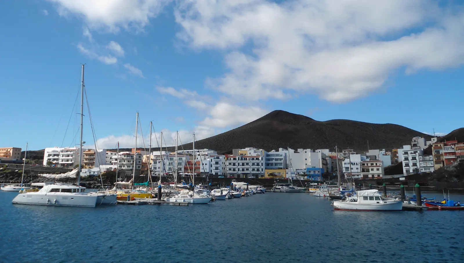 Puerto de La Restinga, en El Hierro, con veleros y barcas de pesca amarrados y el caserío blanco del pueblo al pie de un cono volcánico.