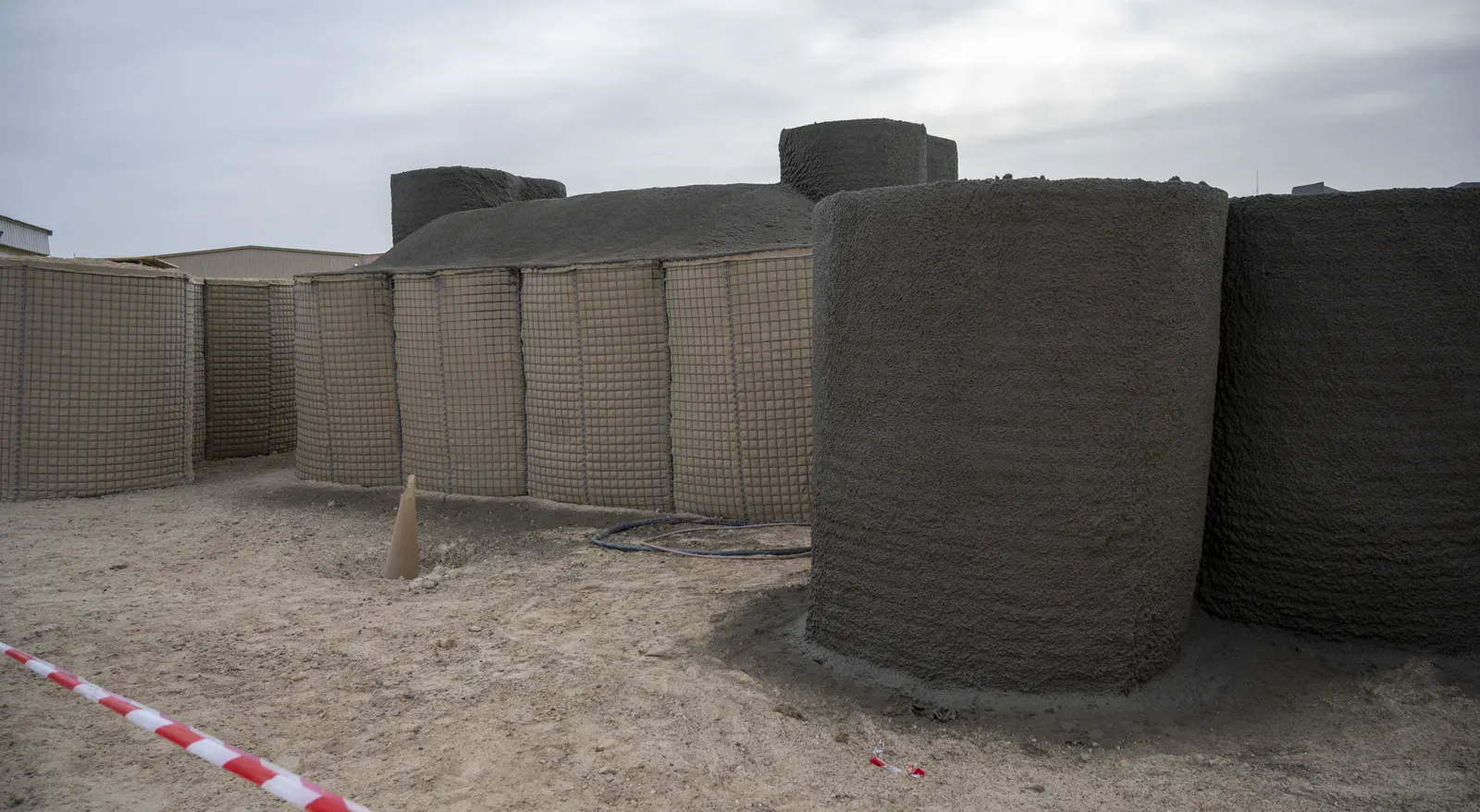 Búnkeres de hormigón y muros de gaviones metálicos rellenos de arena sobre suelo de tierra en la base aérea de Alí al Salem, en Kuwait, con cielo nublado al fondo.