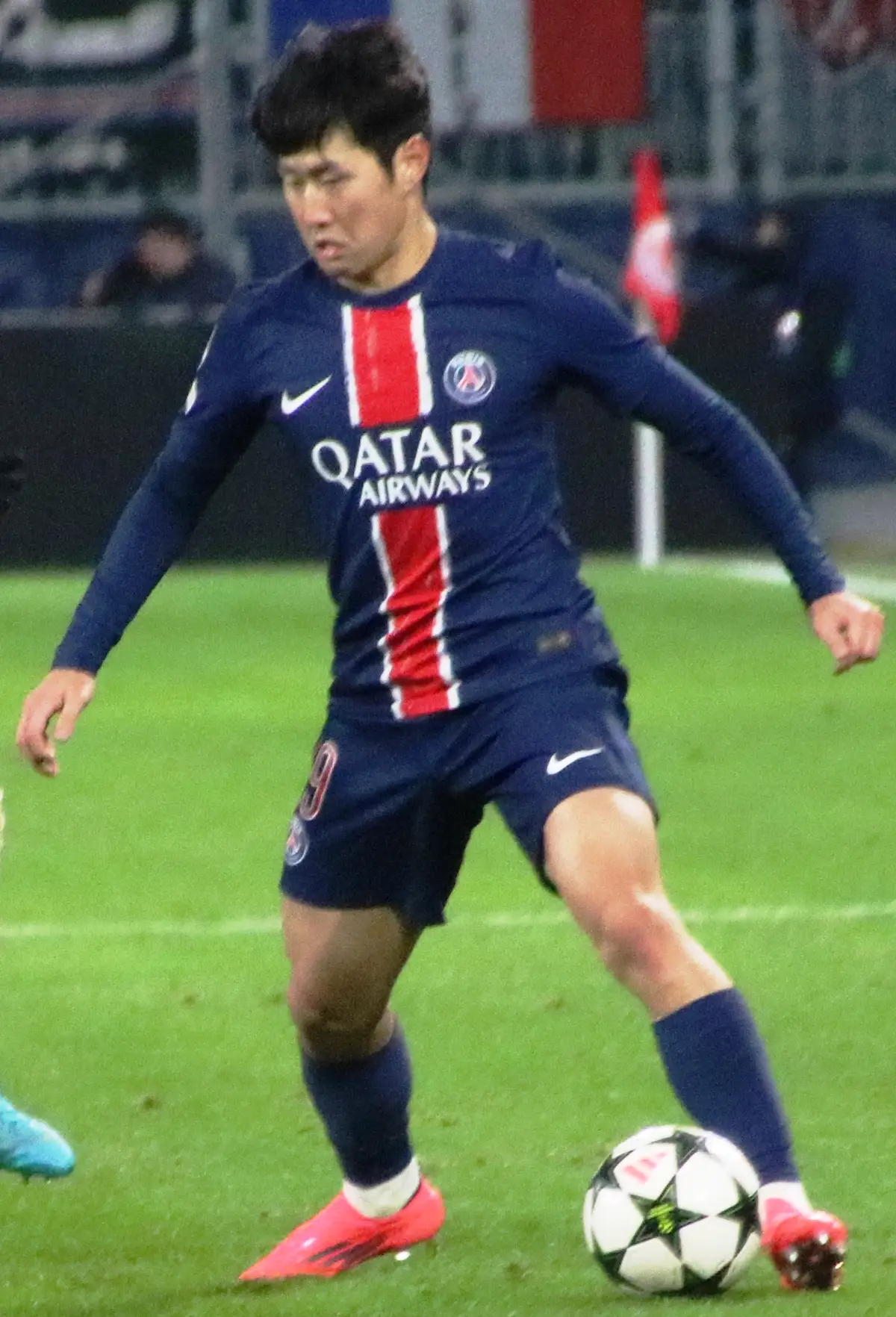 Kang-in Lee, con la camiseta azul y roja del Paris Saint-Germain, conduce el balón con la pierna izquierda sobre el césped durante un partido nocturno