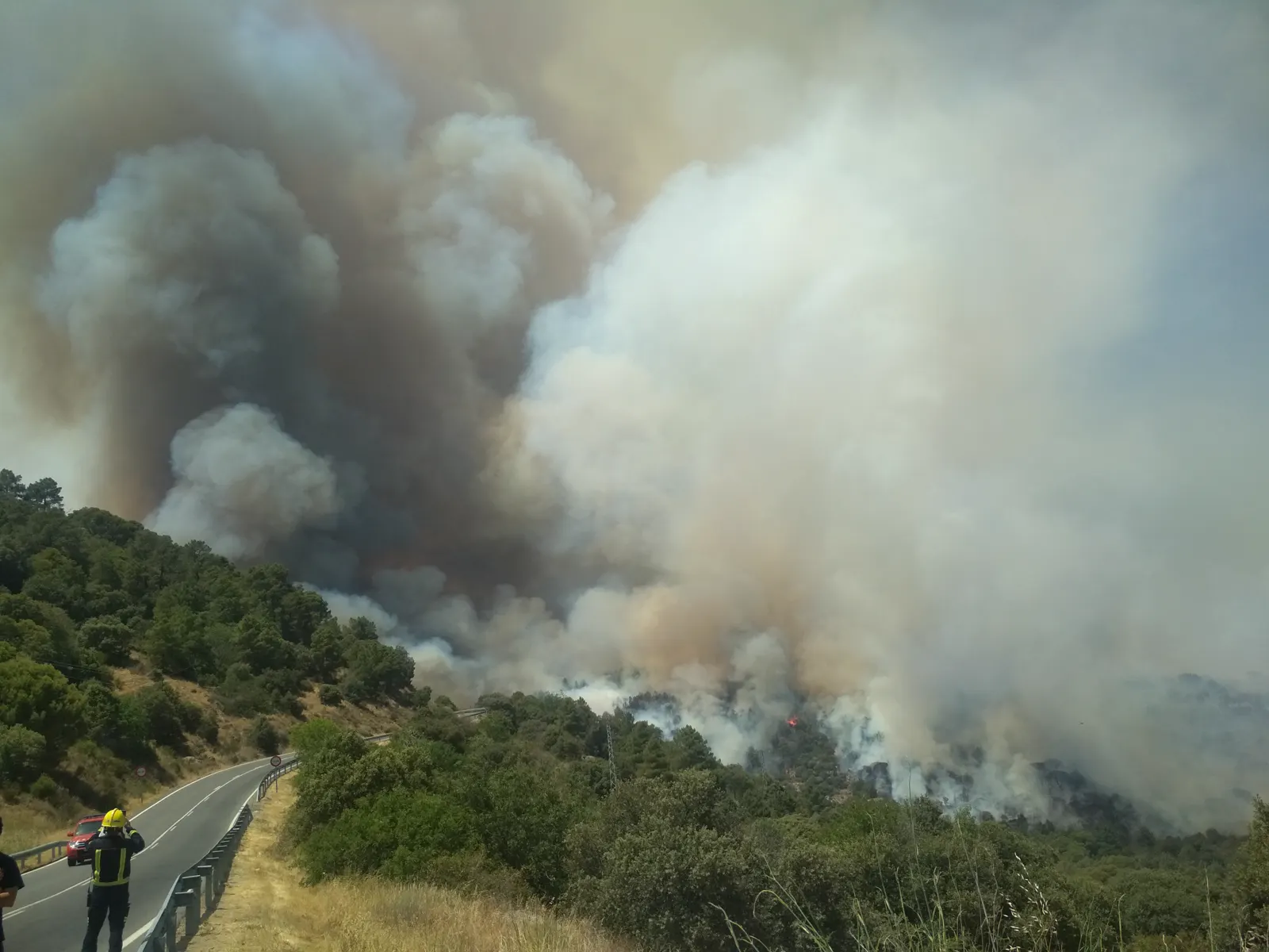 Un bombero con casco amarillo observa desde el arcén de una carretera de montaña una enorme columna de humo gris que sale de una ladera de pinar y encinar; entre el humo se distinguen llamas anaranjadas.