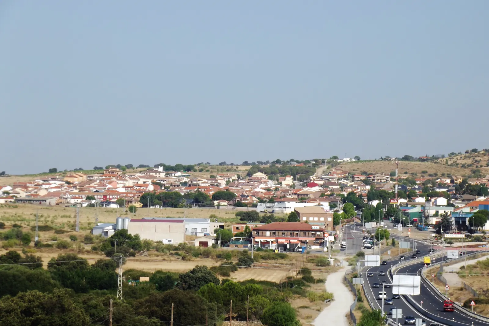 Vista general de Navas del Rey (Madrid): casas de teja roja extendidas sobre una loma de pastos secos y encinar, con naves industriales y una carretera de doble calzada en primer término.