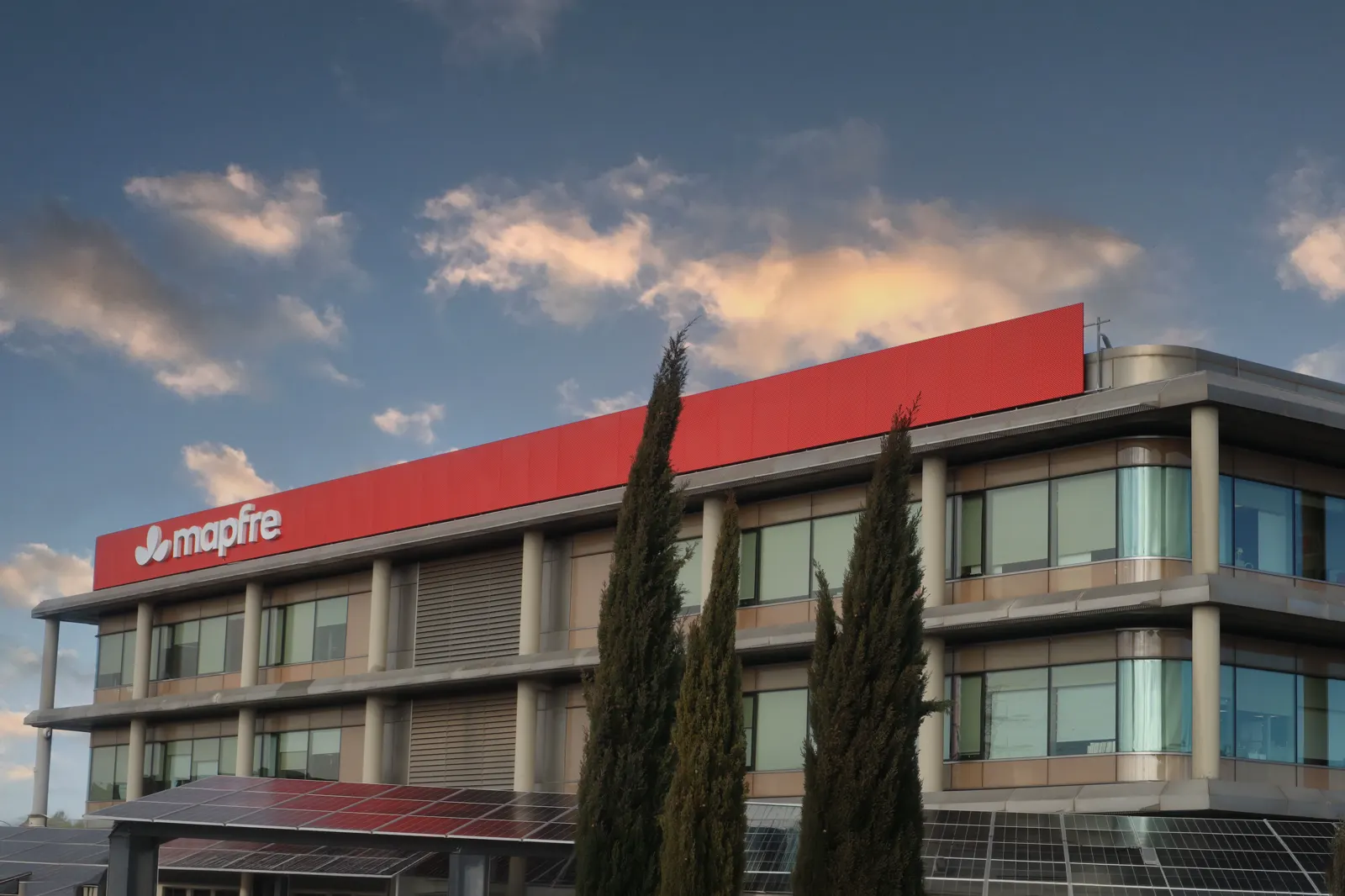 Edificio de oficinas de tres plantas coronado por un cartel rojo con el logotipo de Mapfre, con cipreses delante y placas solares en la marquesina.