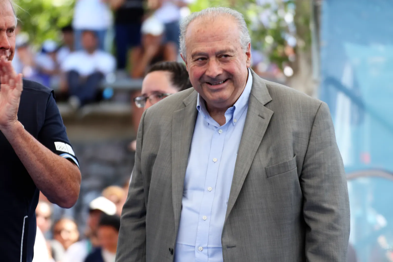 Hombre mayor de pelo cano, con americana gris claro y camisa azul, sonríe durante un acto deportivo al aire libre; a su lado, un hombre aplaude y detrás se ve público en una grada