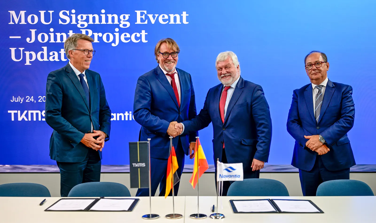 Cuatro directivos trajeados posan tras una mesa con dos carpetas abiertas; los dos del centro se estrechan la mano ante banderas de Alemania y España y un panel azul con la leyenda «MoU Signing Event – Joint Project Update, July 24, 2026» y los logotipos de TKMS y Navantia.