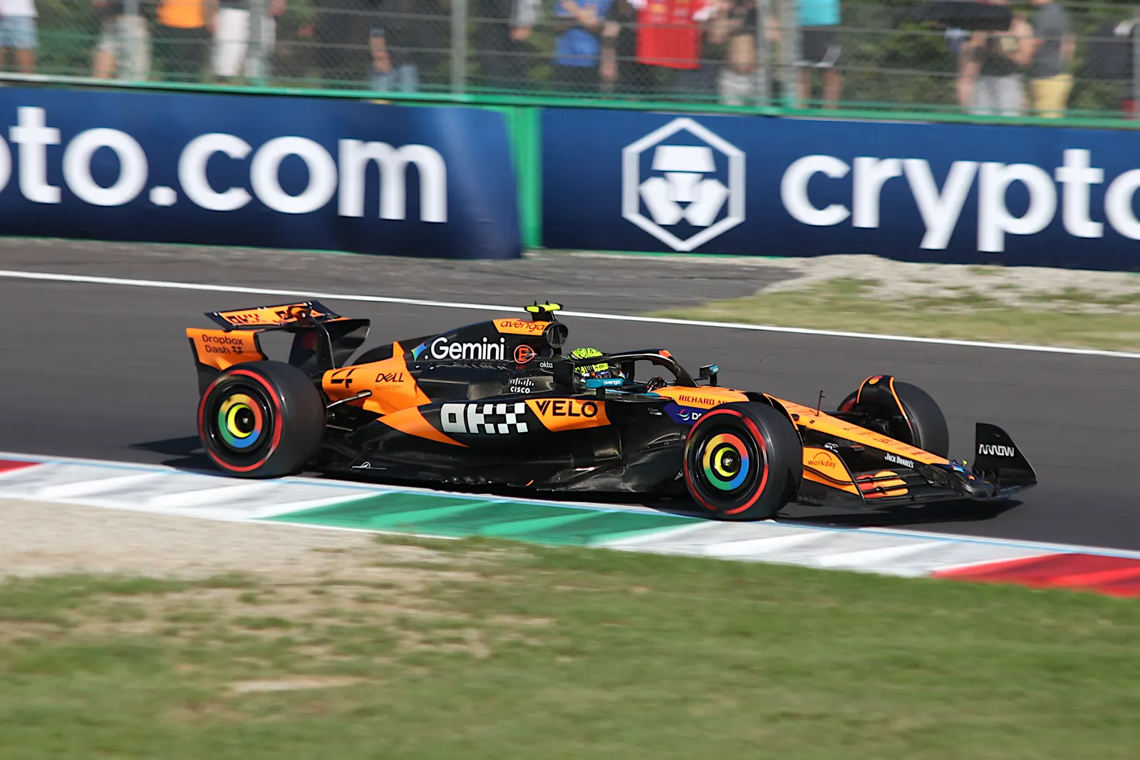 Lando Norris, con casco amarillo y flúor, al volante de su McLaren naranja y negro en una recta de circuito.
