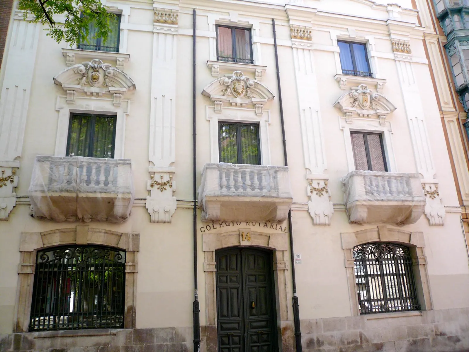 Fachada clara de un edificio señorial con tres balcones de balaustrada de piedra, escudos esculpidos sobre las ventanas y la inscripción «Colegio Notarial» tallada sobre el portón de madera del número 14.