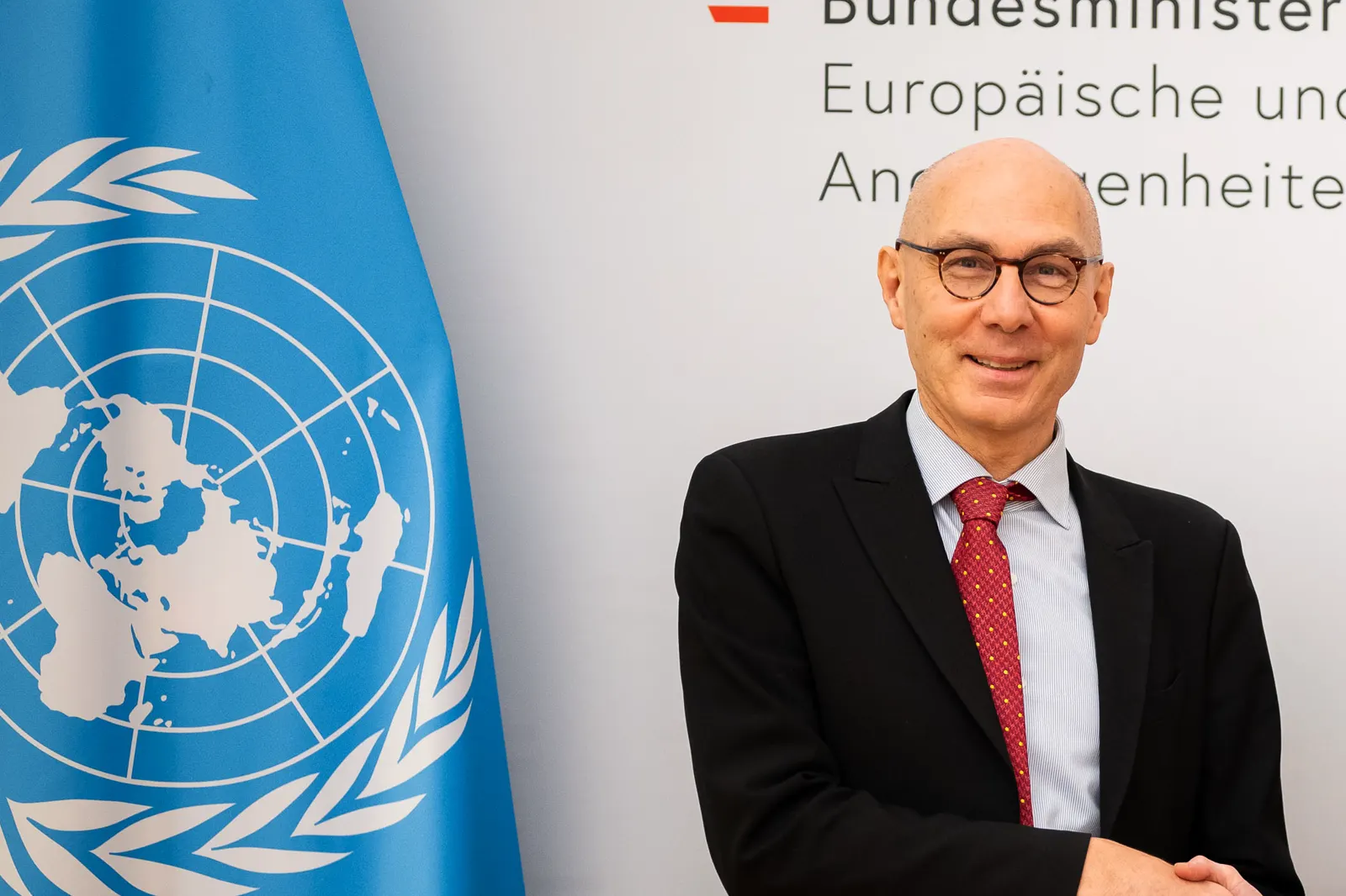 Volker Türk, con traje oscuro, gafas y corbata roja, posa sonriente junto a una bandera azul de Naciones Unidas ante un panel con el rótulo del Ministerio de Exteriores austriaco