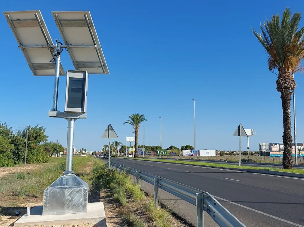 Radar fijo de velocidad con dos placas solares, instalado junto a una carretera de doble sentido con palmeras y cielo despejado.