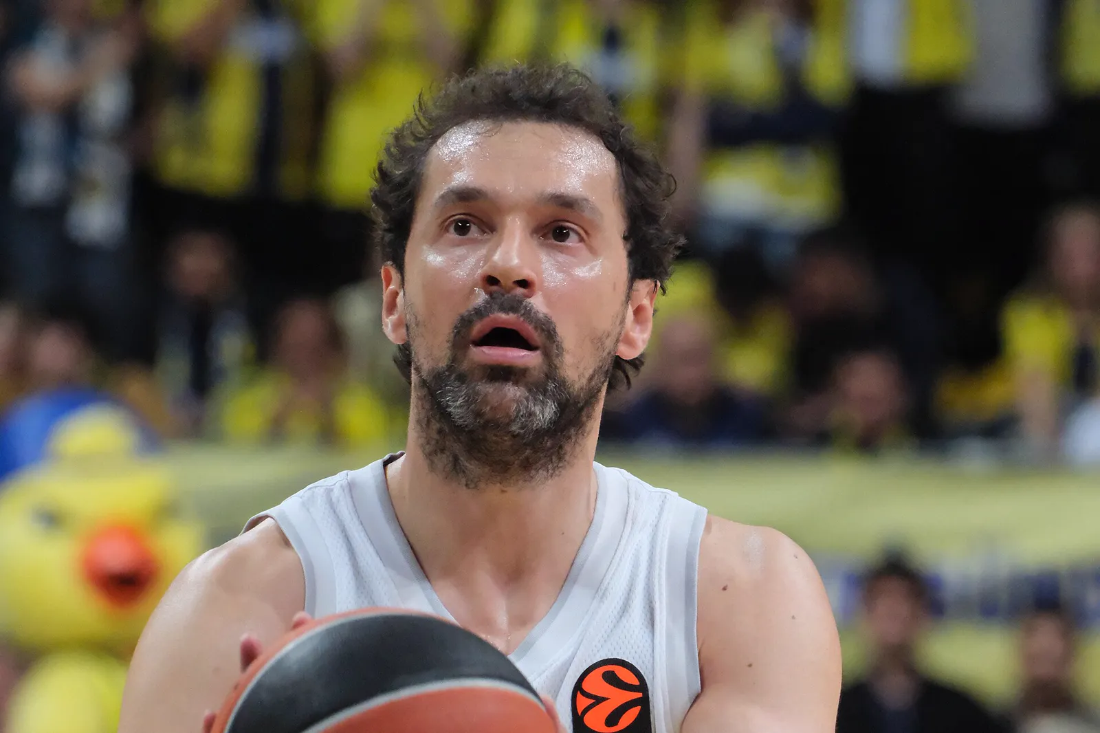 Sergio Llull, con la camiseta blanca del Real Madrid, sujeta el balón con las dos manos y mira hacia el aro antes de lanzar un tiro libre; al fondo, la grada del pabellón vestida de amarillo