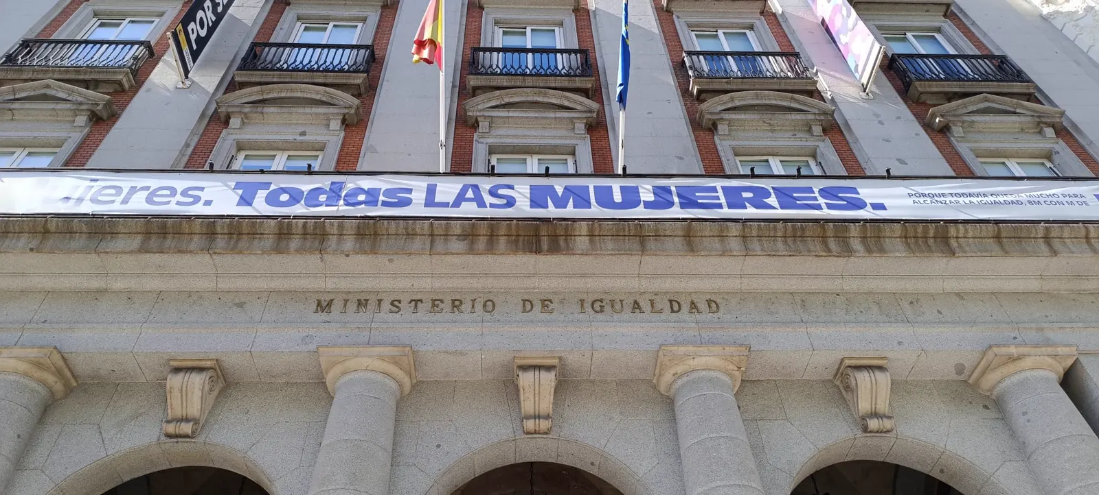 Fachada del Ministerio de Igualdad en Madrid vista desde abajo: sobre la entrada porticada de columnas, la inscripción en piedra «Ministerio de Igualdad» y, encima, una pancarta blanca con el lema «Todas las mujeres» junto a una bandera de España y otra azul.
