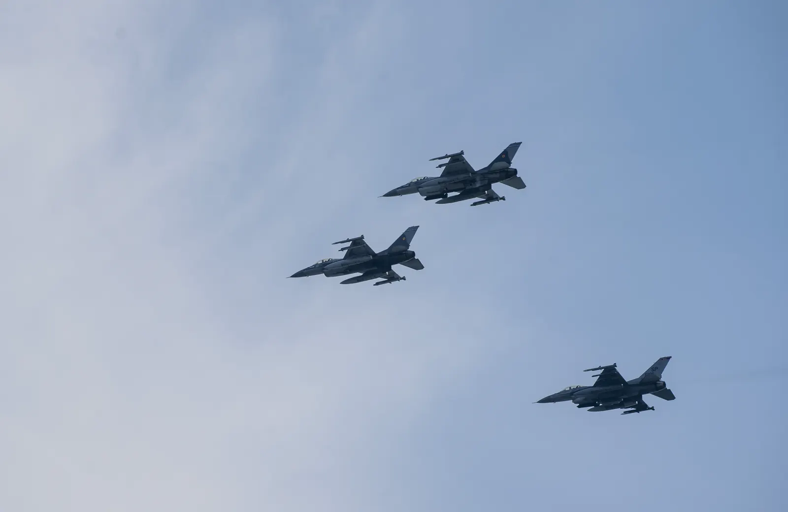 Tres cazas F-16 vuelan en formación escalonada sobre un cielo azul con bruma; los dos de arriba llevan la bandera rumana en la deriva y el tercero, el código de cola estadounidense SP.