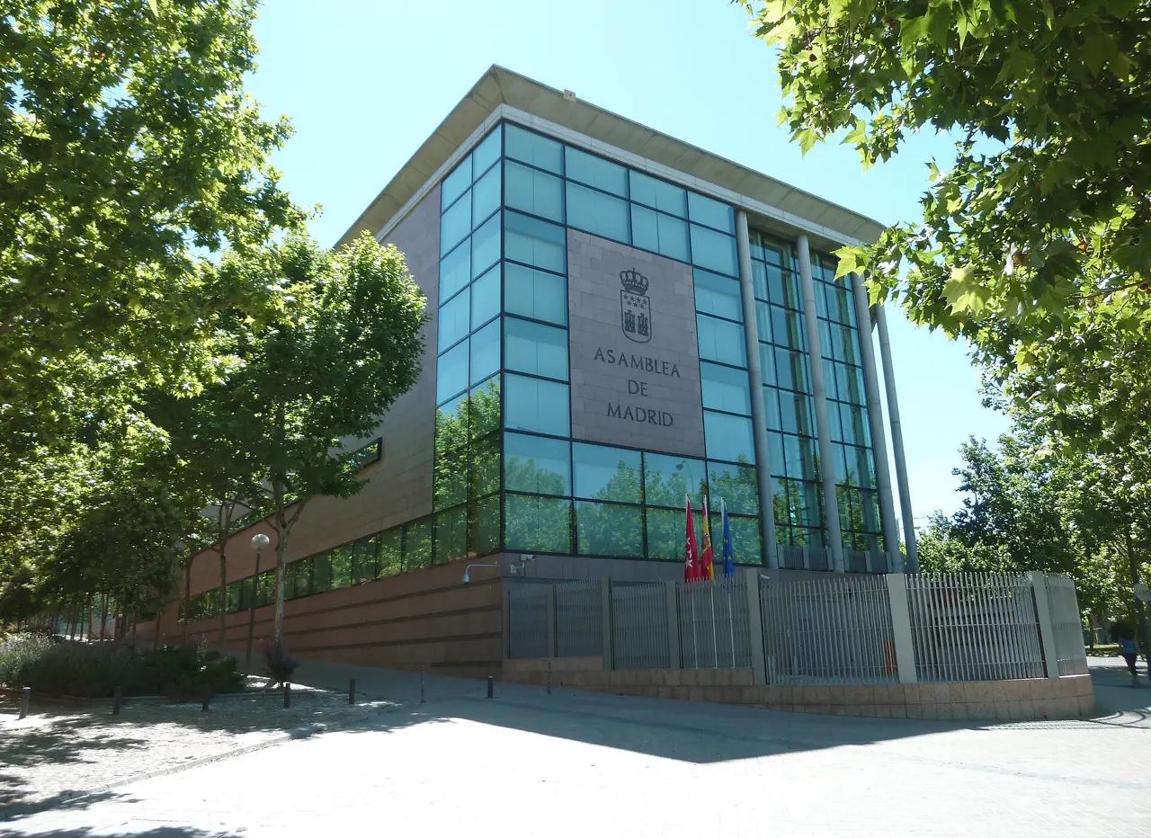 Fachada acristalada y de piedra de la sede de la Asamblea de Madrid, con el escudo de la Comunidad y el rótulo «Asamblea de Madrid» sobre el paño central, banderas de la Comunidad, España y la Unión Europea ante la entrada y árboles a ambos lados.