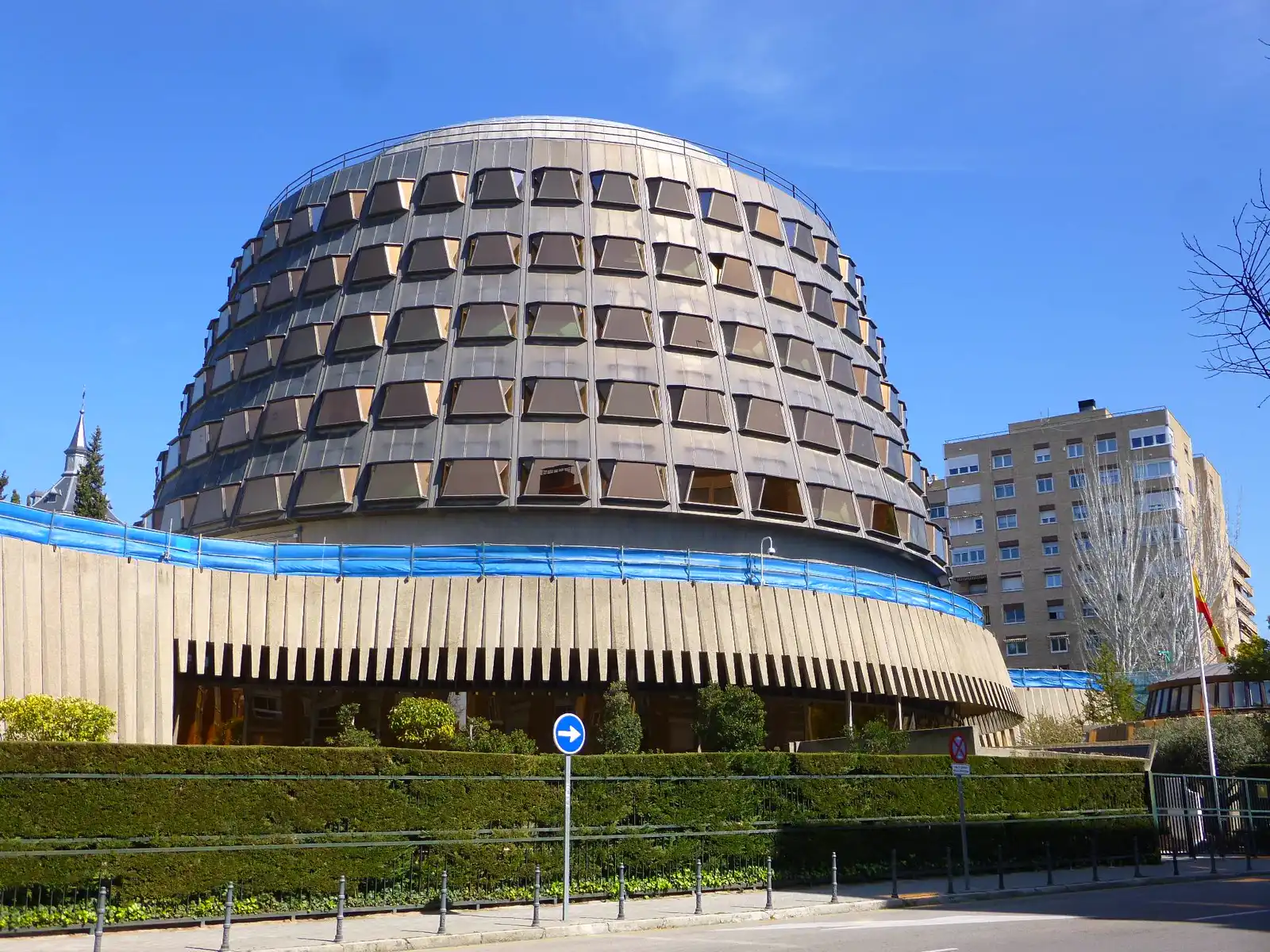Edificio circular del Tribunal Constitucional en Madrid, con fachada de ventanales oscuros inclinados sobre un zócalo de hormigón, un seto y una verja en primer plano y la bandera de España en un mástil a la derecha.