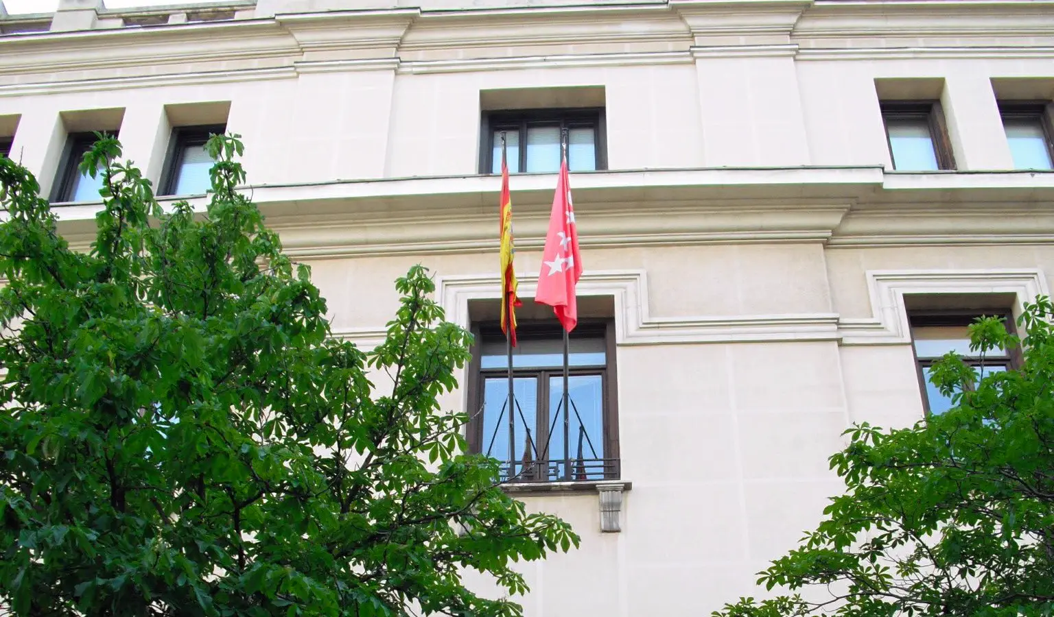Fachada de piedra clara del edificio del Tribunal Superior de Justicia de Madrid, con las banderas de España y de la Comunidad de Madrid colgadas de un balcón central y las copas de dos castaños en primer plano.
