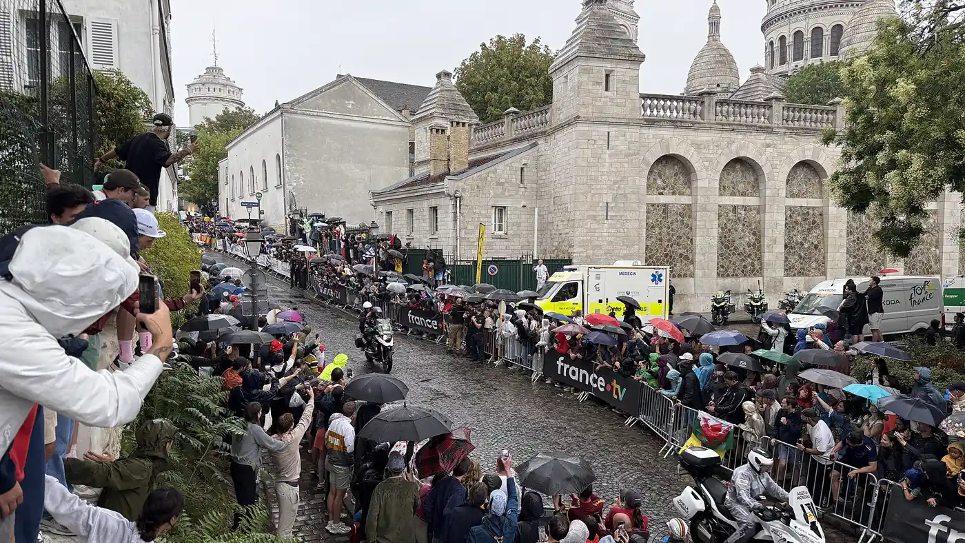 Una calle empedrada y en cuesta de Montmartre, bajo la lluvia, abarrotada de público con paraguas tras las vallas; una moto de la organización sube por el centro de la calzada y al fondo asoman las cúpulas de la basílica del Sacré-Coeur.