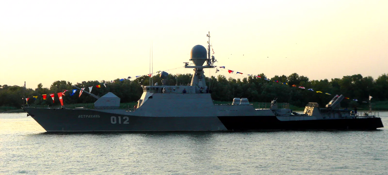 Buque de guerra gris con el nombre АСТРАХАНЬ y el número 012 en el casco, engalanado con banderas de señales de colores, navegando sobre agua en calma con una orilla arbolada al fondo.