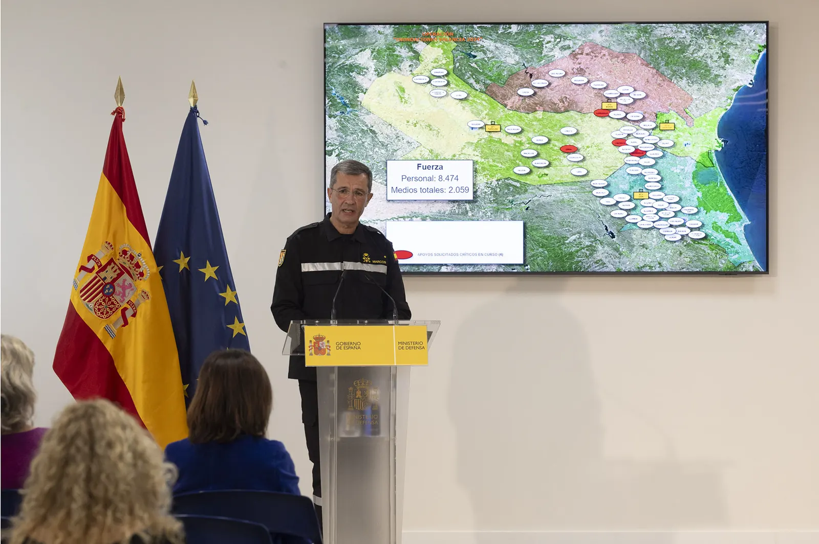 El jefe de la UME, Francisco Javier Marcos Izquierdo, con mono de trabajo negro, habla en un atril del Ministerio de Defensa junto a las banderas de España y de la Unión Europea; detrás, una pantalla con un mapa de la operación y el recuento de personal y medios desplegados.