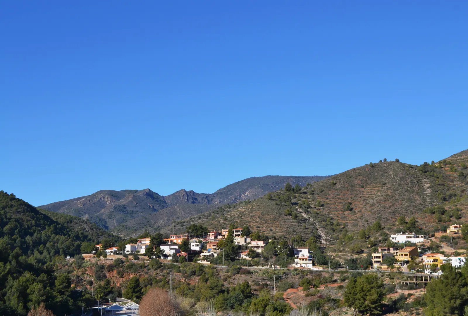 Casas dispersas de una urbanización en una ladera cubierta de pinar, con las montañas de la Serra d'Espadà al fondo bajo un cielo despejado.