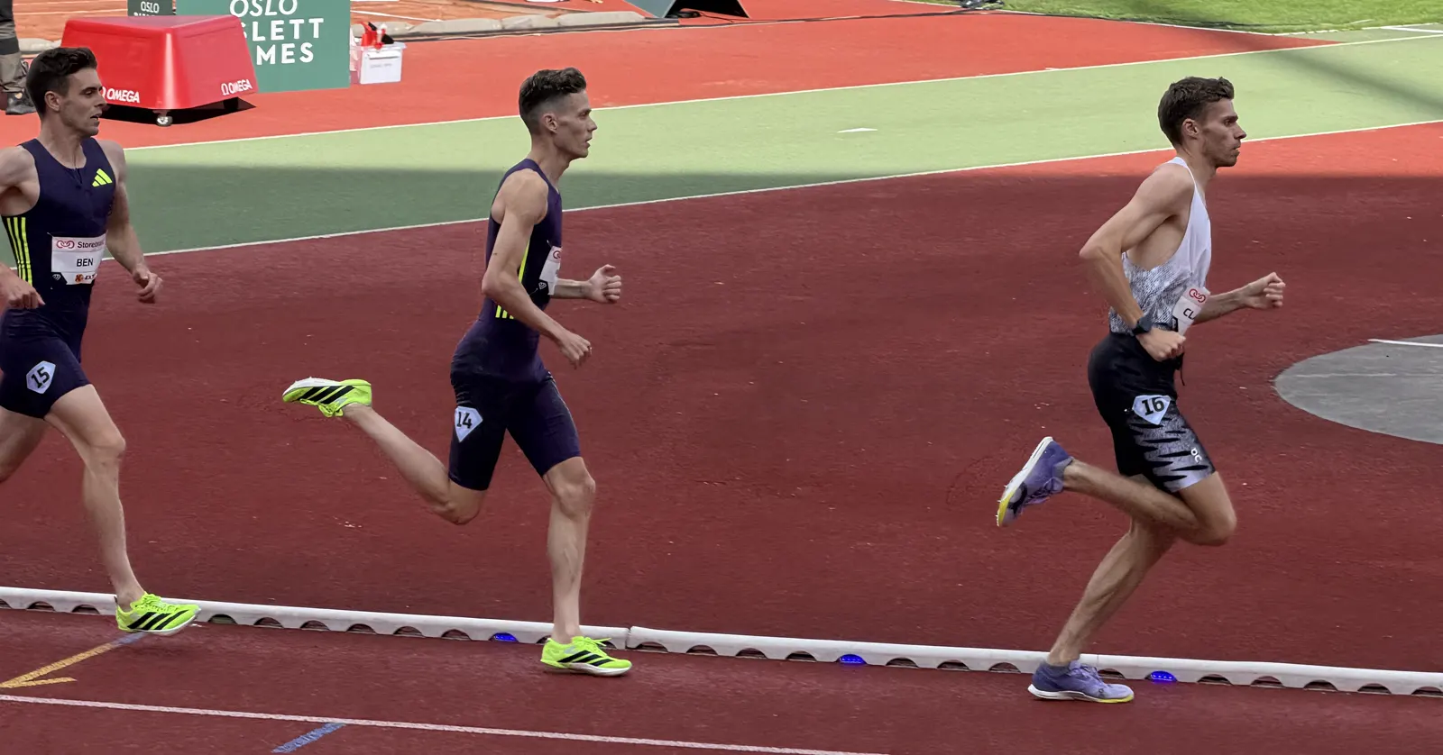 Tres atletas corren en fila por la pista de tartán de un estadio; el que va último, a la izquierda, lleva un maillot azul oscuro con el dorsal con el nombre BEN y el número 15.