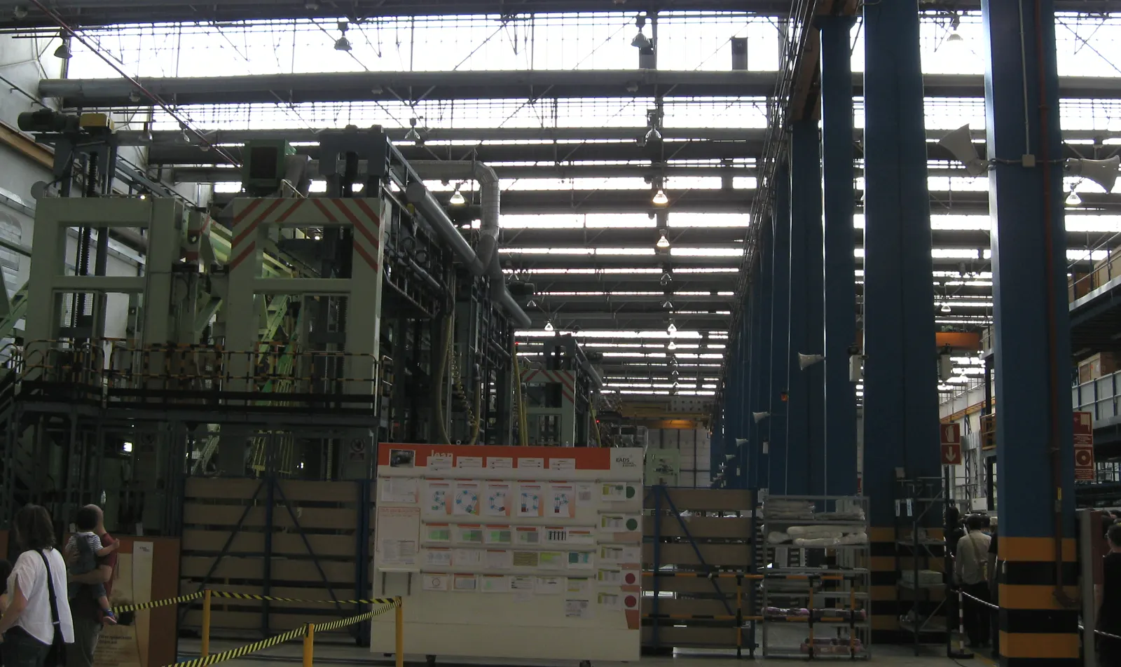 Interior de una nave industrial de gran altura con estructura metálica y lucernarios en el techo, utillaje verde de gran tamaño a la izquierda, pilares azules, estanterías con material y paneles informativos junto a una zona acordonada.