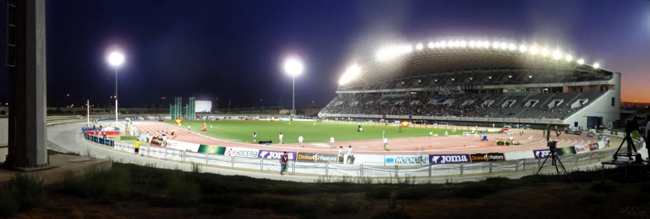 Panorámica nocturna del Estadio Ciudad de Málaga iluminado por focos, con la pista de tartán, atletas sobre el césped interior, vallas publicitarias y público en la grada cubierta.