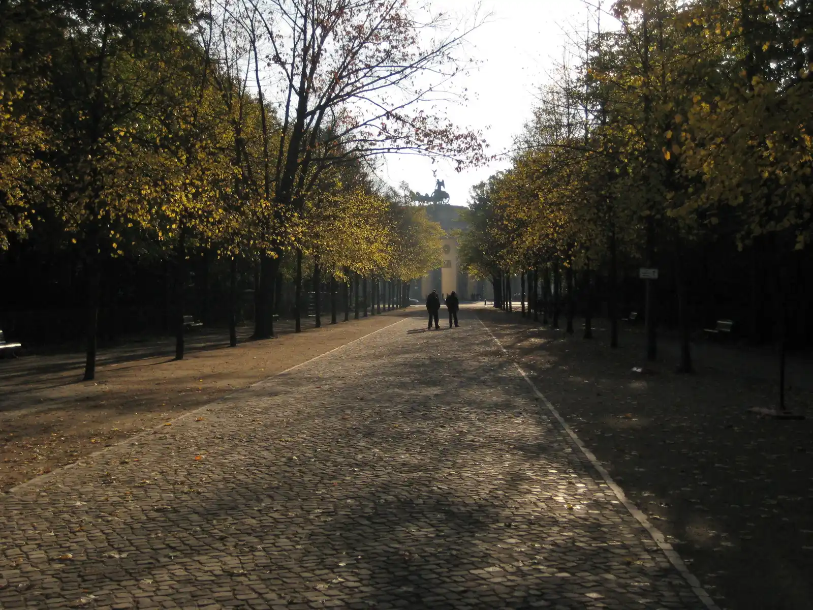 Paseo empedrado y arbolado del Ahornsteig, en el Gran Tiergarten de Berlín, con la cuadriga de la Puerta de Brandeburgo al fondo.