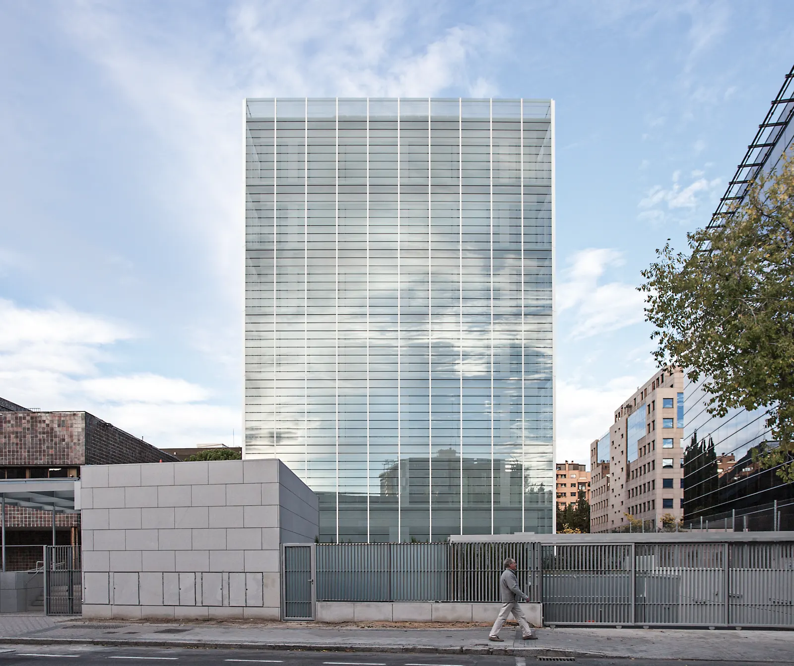 Edificio de oficinas de fachada acristalada y planta rectangular, cerrado por una verja metálica; un hombre camina por la acera en primer término bajo un cielo azul con nubes.