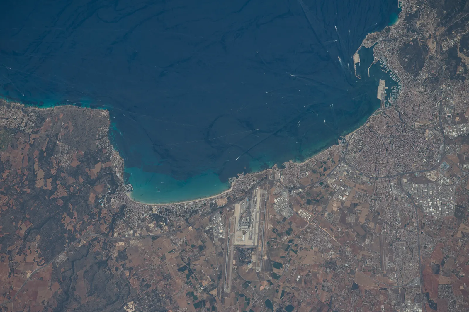 Fotografía tomada desde la Estación Espacial Internacional de la bahía de Palma, en el sur de Mallorca: el mar azul oscuro ocupa la mitad superior, con la ciudad y el puerto de Palma a la derecha, la playa de arena clara en el centro y las pistas del aeropuerto de Son Sant Joan en el interior.