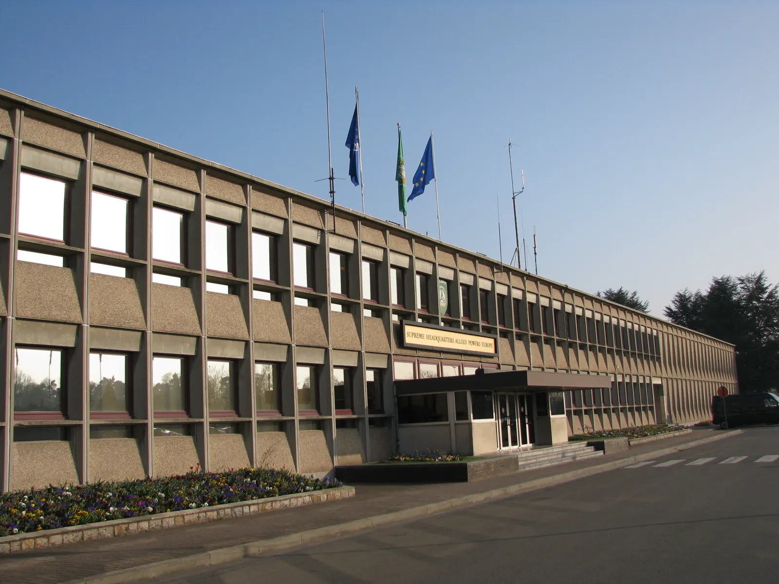 Fachada alargada del edificio principal del cuartel general del SHAPE, con el rótulo «Supreme Headquarters Allied Powers Europe» sobre la entrada y tres banderas izadas en el tejado.