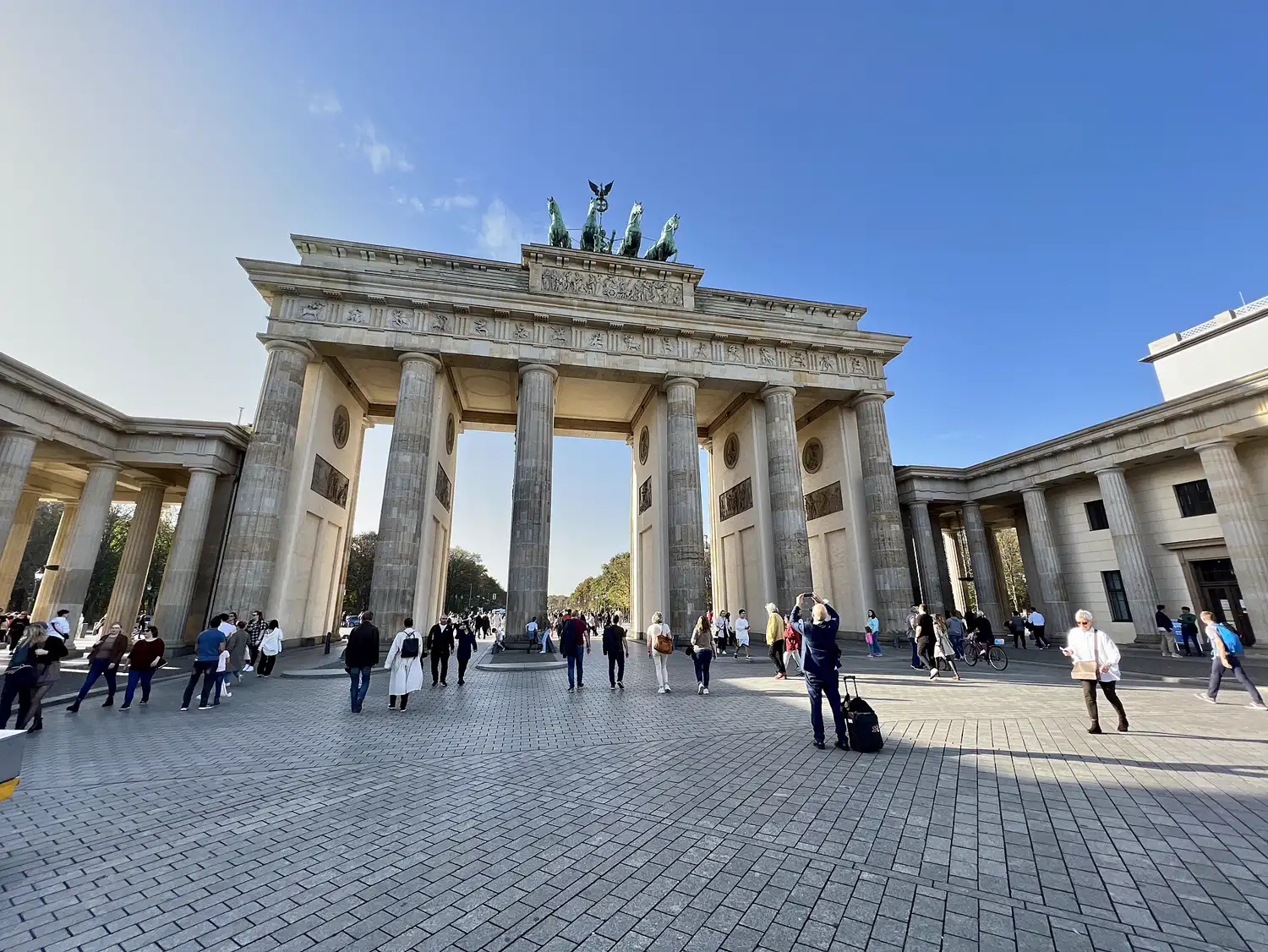Vista de la Puerta de Brandeburgo desde la Pariser Platz de Berlín, con viandantes caminando por el empedrado y el arbolado del Tiergarten visible al fondo entre las columnas.