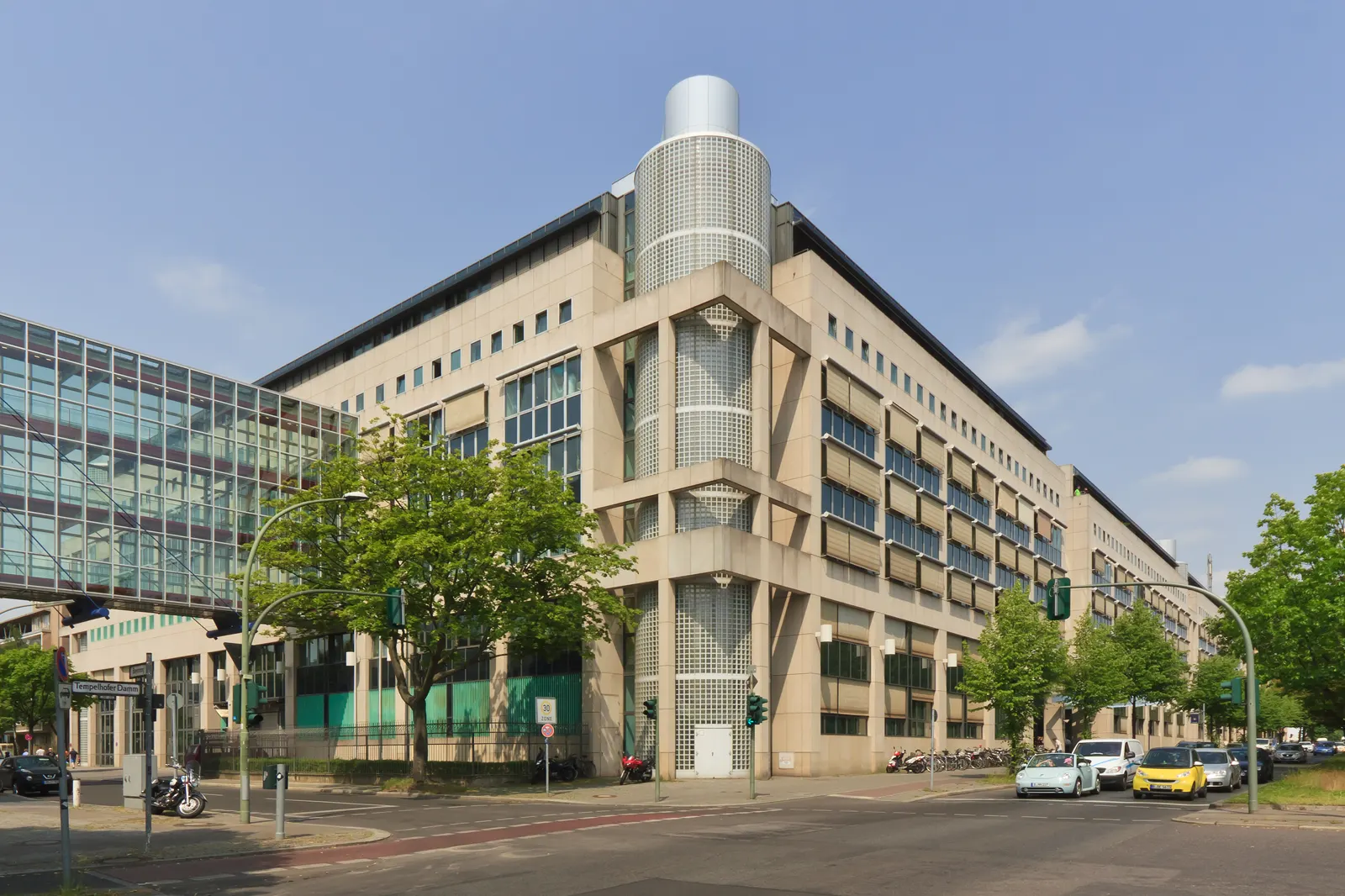 Edificio moderno de piedra clara y vidrio en una esquina del Tempelhofer Damm de Berlín, sede de la Jefatura de Policía, con árboles y tráfico delante.